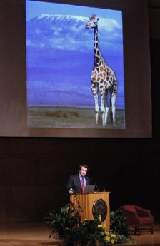 Greg Mortenson presenting with a slide of a giraffe in the background